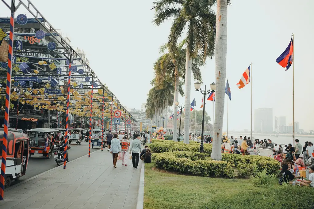 Bustling scene along Phnom Penh riverside with tuk-tuks, people, and flags creating a lively atmosphere.