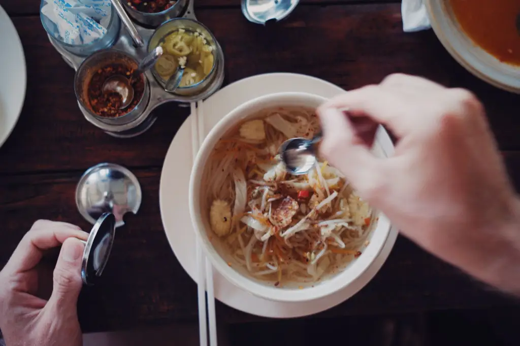 Top-view shot of hands preparing to eat a flavorful Asian noodle soup with various spices and chopsticks.