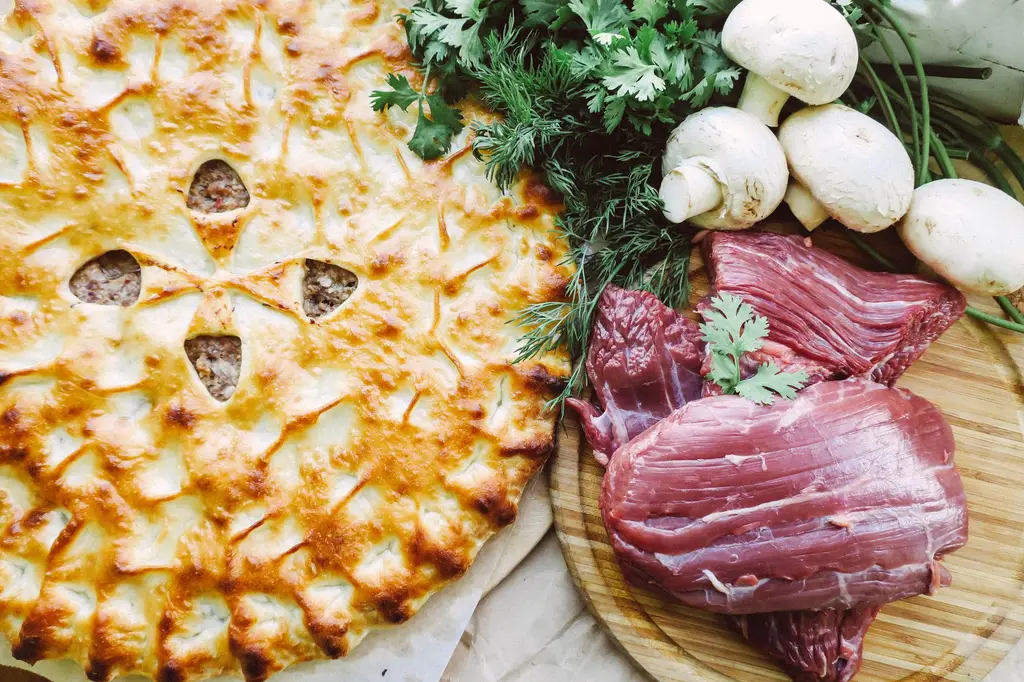 Top view of a meat pie with raw beef, mushrooms, and herbs on a cutting board.