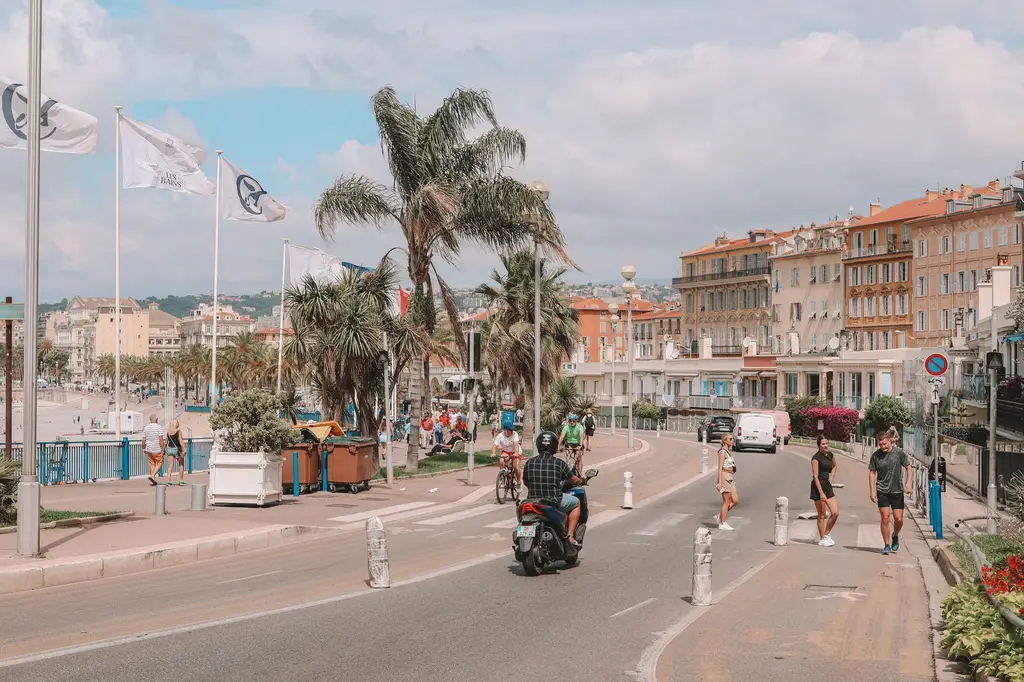 Pedestrians and scooters on bustling Promenade des Anglais in Nice, France, with palm trees and coastal buildings.