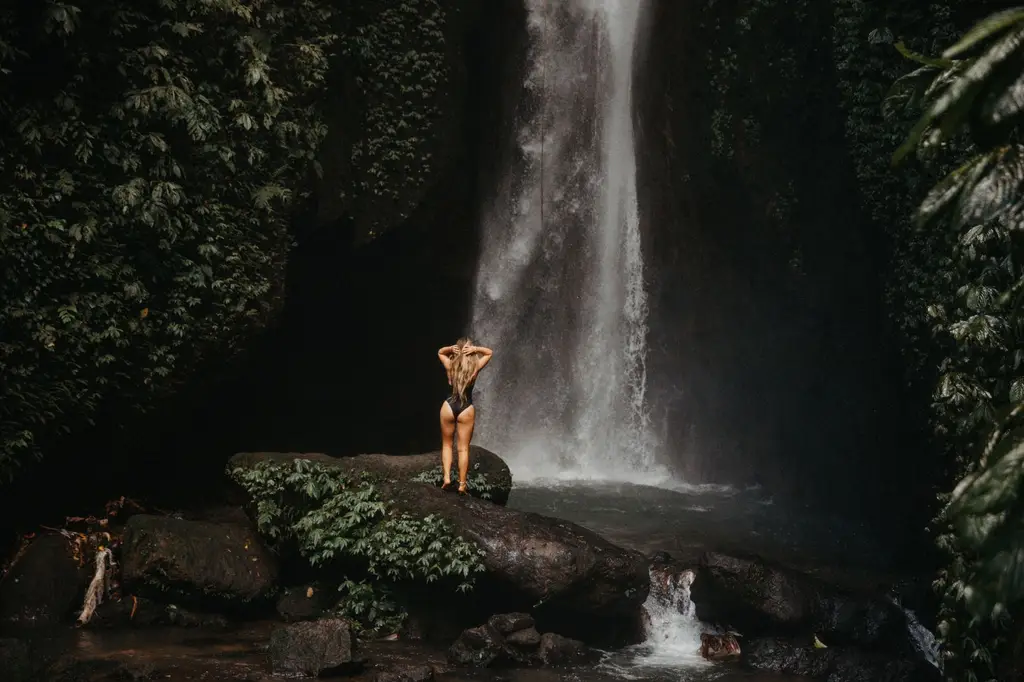 A woman stands by a serene waterfall in Bali's vibrant rainforest, embracing nature's beauty.