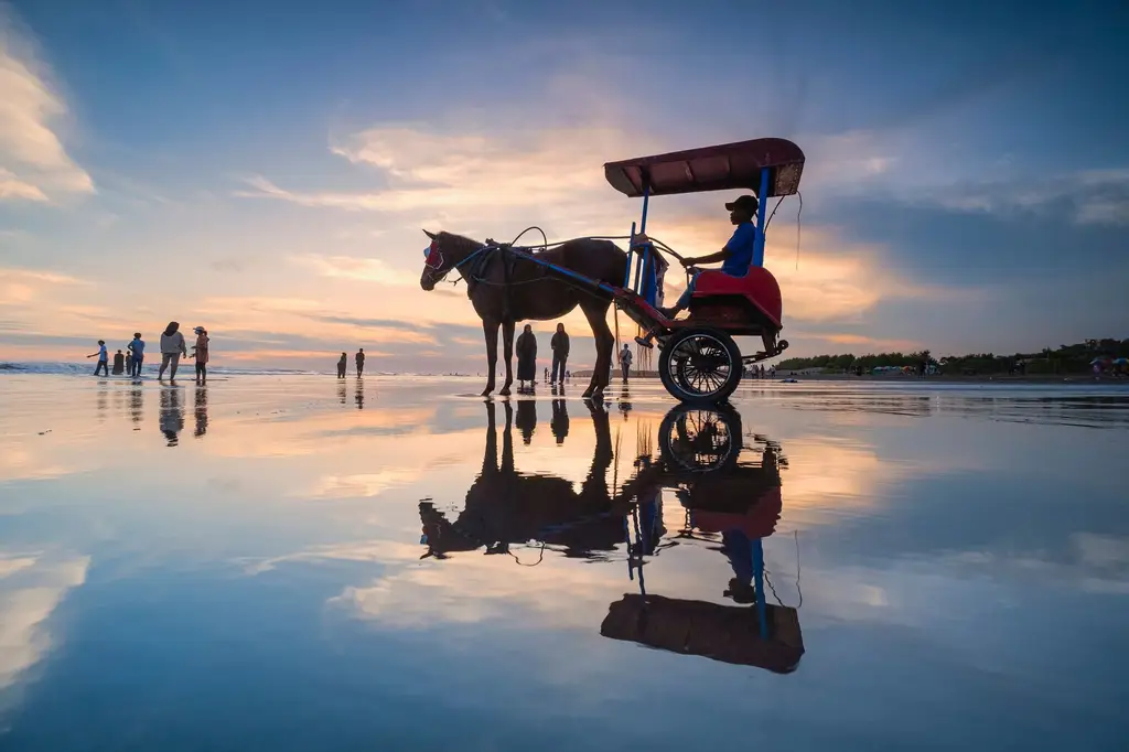 A horse-drawn carriage on Parangtritis Beach at sunset creates a serene reflection on the wet sand.