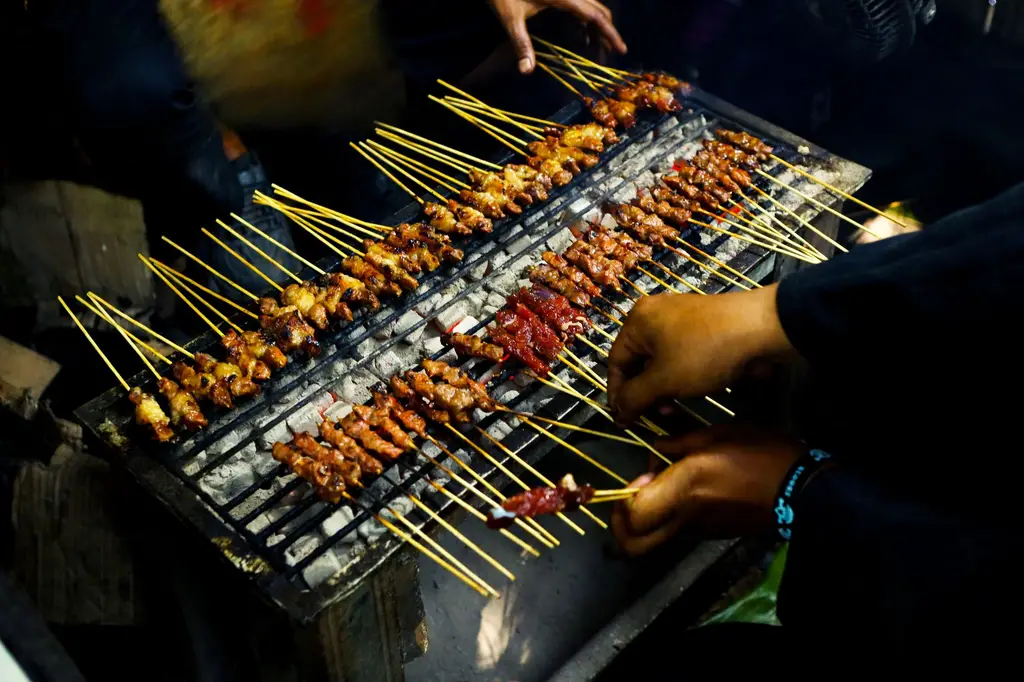 Juicy chicken and beef sate skewers grilling over charcoal at a bustling Indonesian night market.