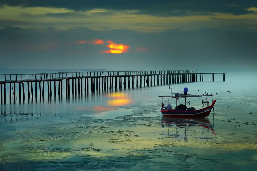 Serene sunrise view with boat and pier in George Town, Malaysia, reflecting vibrant colors on the sea.