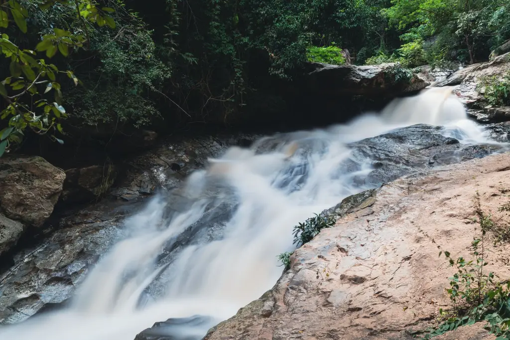 A tranquil waterfall cascades over rocks in the lush greenery of Chiang Mai, Thailand, captured in long exposure.