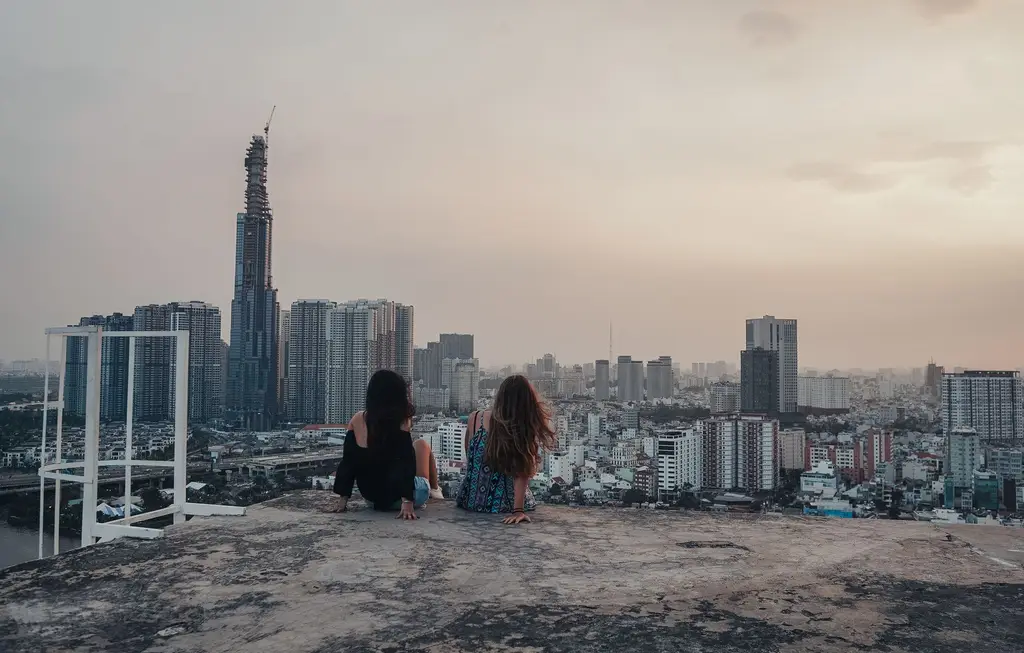 Two women sitting on a rooftop overlooking the Ho Chi Minh City skyline during sunset.